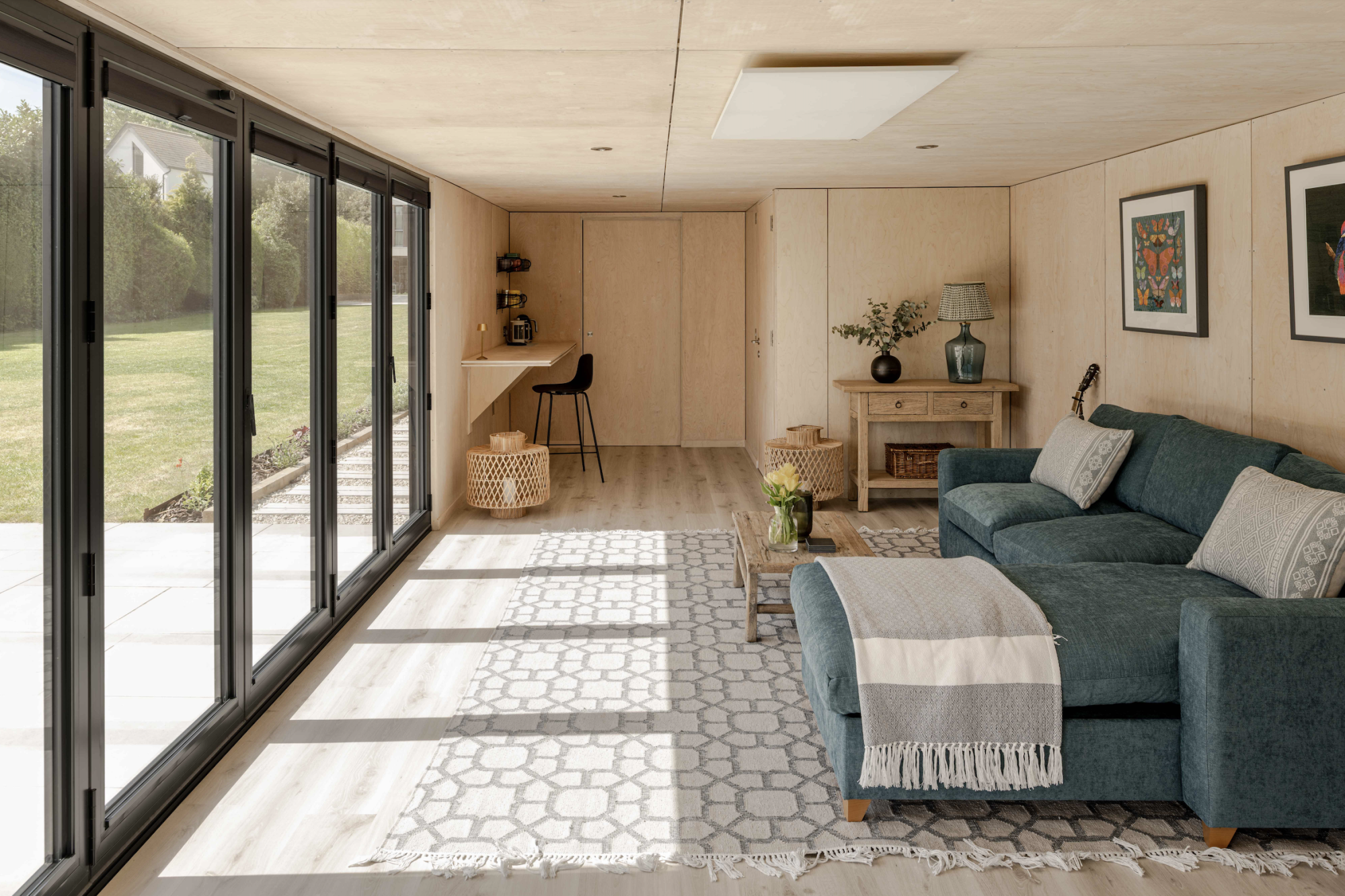 Interior view of a spacious garden room featuring plywood walls, a teal sectional sofa on a geometric rug, and large bifold glass doors overlooking a manicured lawn.